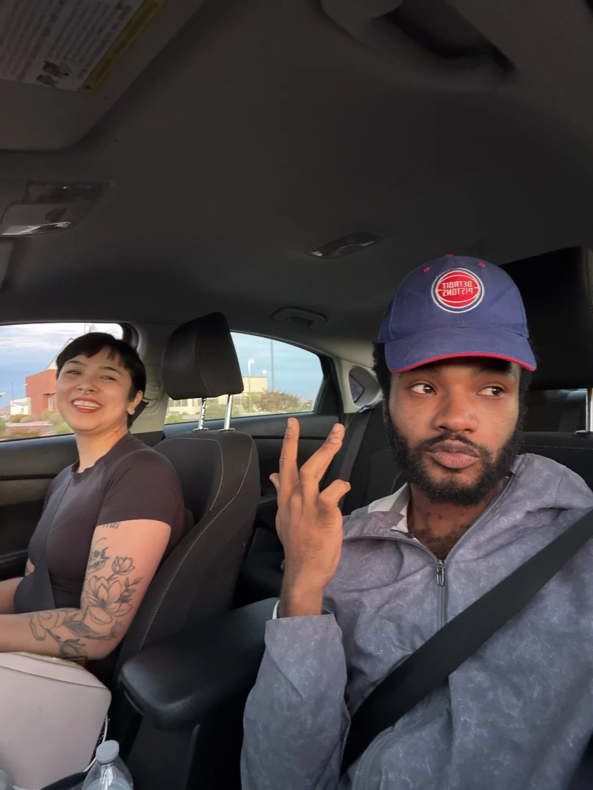 A selfie with a friend in the car, wearing a Detroit Pistons cap and holding up a peace sign.